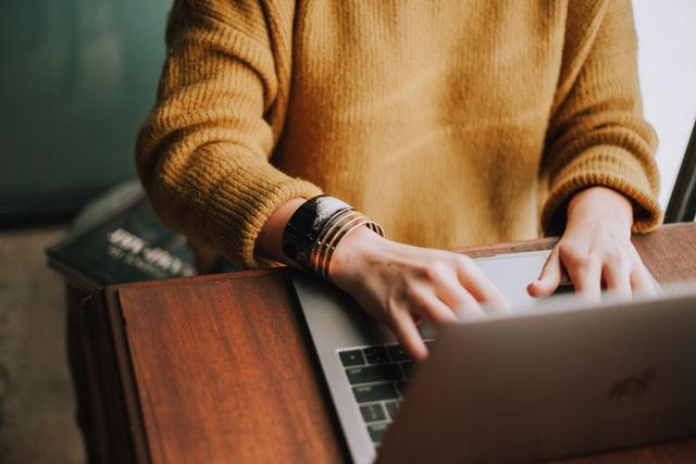 Woman's torso in yellow jumper sits typing on a laptop